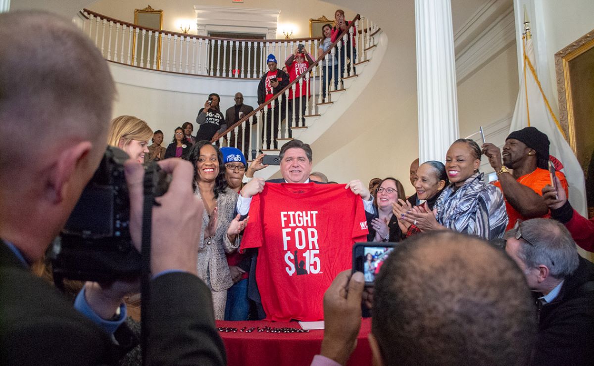 JB Pritzker holds up Fight for $15 t-shirt at the Illinois State Capitol