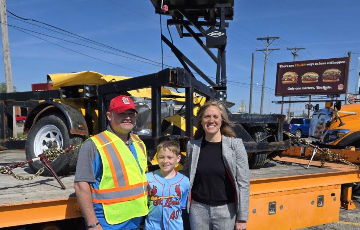 IDOT road maintainer, Steve Wagoner, his son, Hoyt and acting secretary IDOT, Gia Biagi stand in front of a piece of road equipment destroyed by a speeding semi.