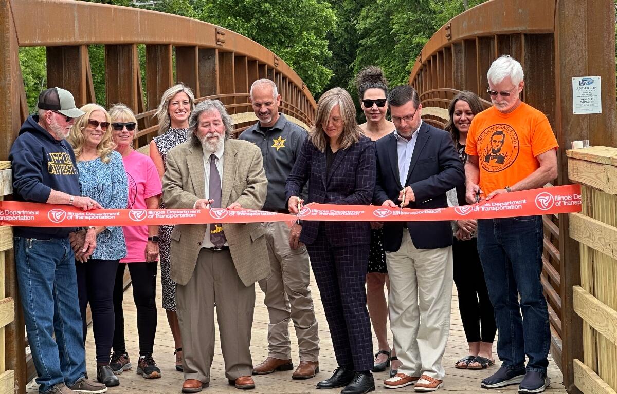 Acting Illinois Department of Transportation Secretary Gia Biagi, towards center wearing dark clothing, helps cut the ribbon on the new Lincoln Prairie Trail Bridge.