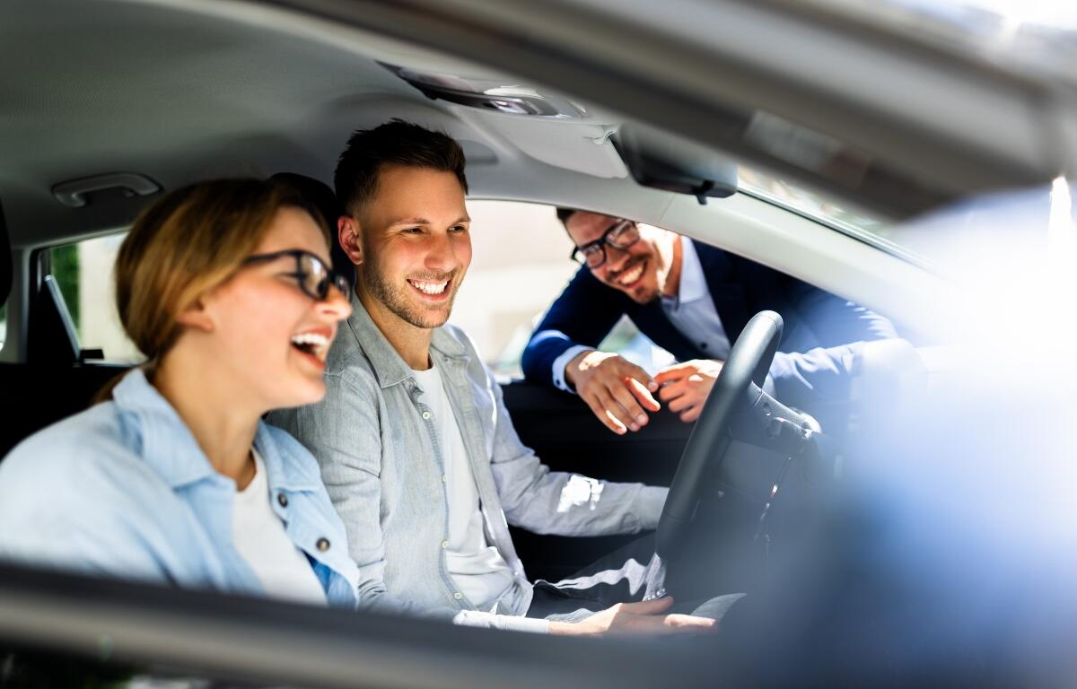 Professional Car Salesman Excitedly Showcases Sleek New Vehicle to Young Couple Inside Dealership, Hispanic Woman Holding Steering Wheel Smiling.