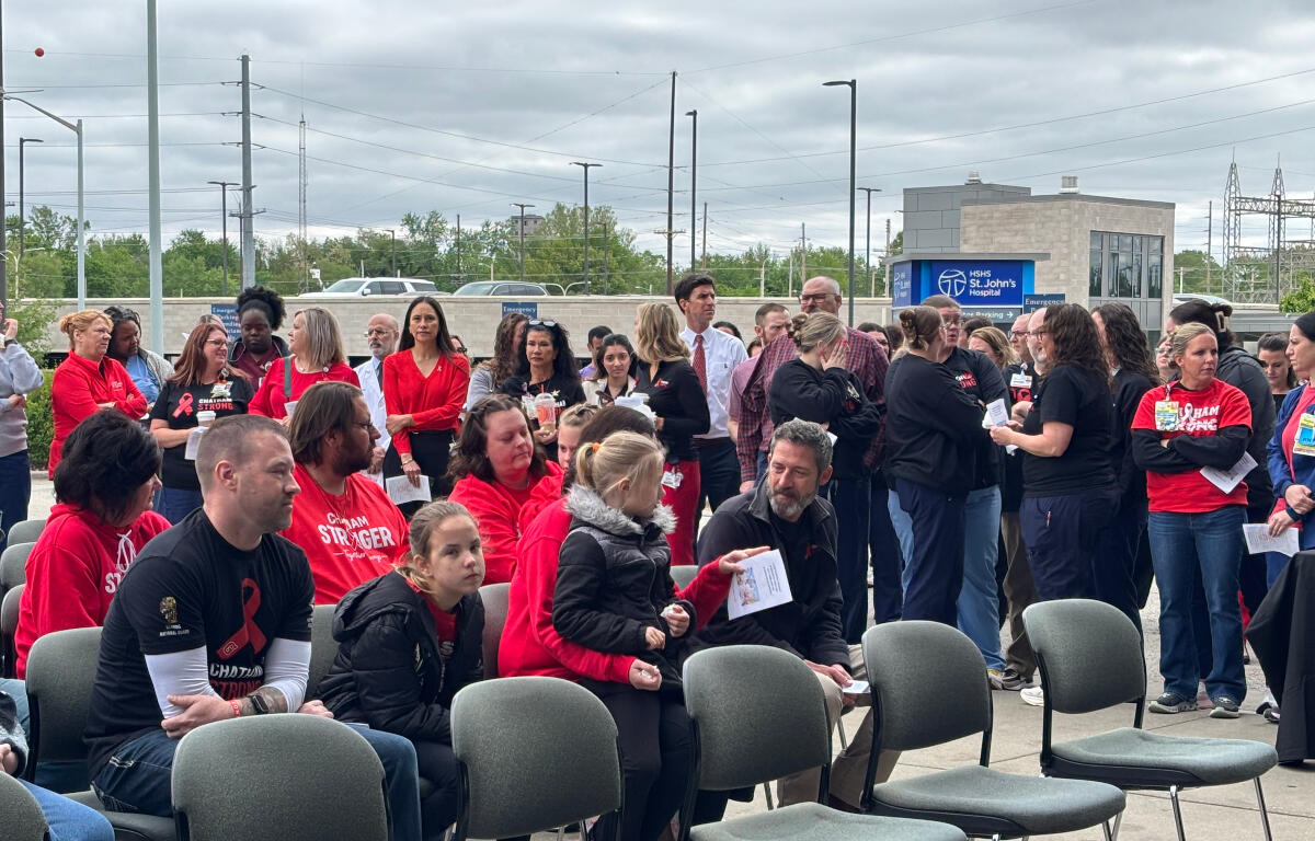 Outdoor gathering of people, many in red shirts, seated and standing near a hospital entrance backdrop.