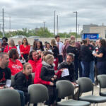 Outdoor gathering of people, many in red shirts, seated and standing near a hospital entrance backdrop.