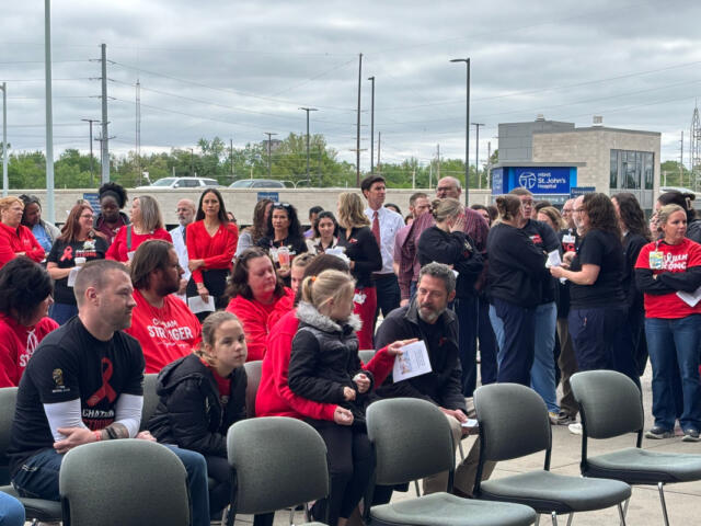 Outdoor gathering of people, many in red shirts, seated and standing near a hospital entrance backdrop.
