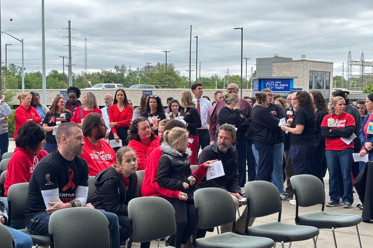 Outdoor gathering of people, many in red shirts, seated and standing near a hospital entrance backdrop.