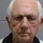 Close-up of an elderly man with short white hair and a neutral, serious expression against a gray wall backdrop.