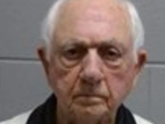 Close-up of an elderly man with short white hair and a neutral, serious expression against a gray wall backdrop.