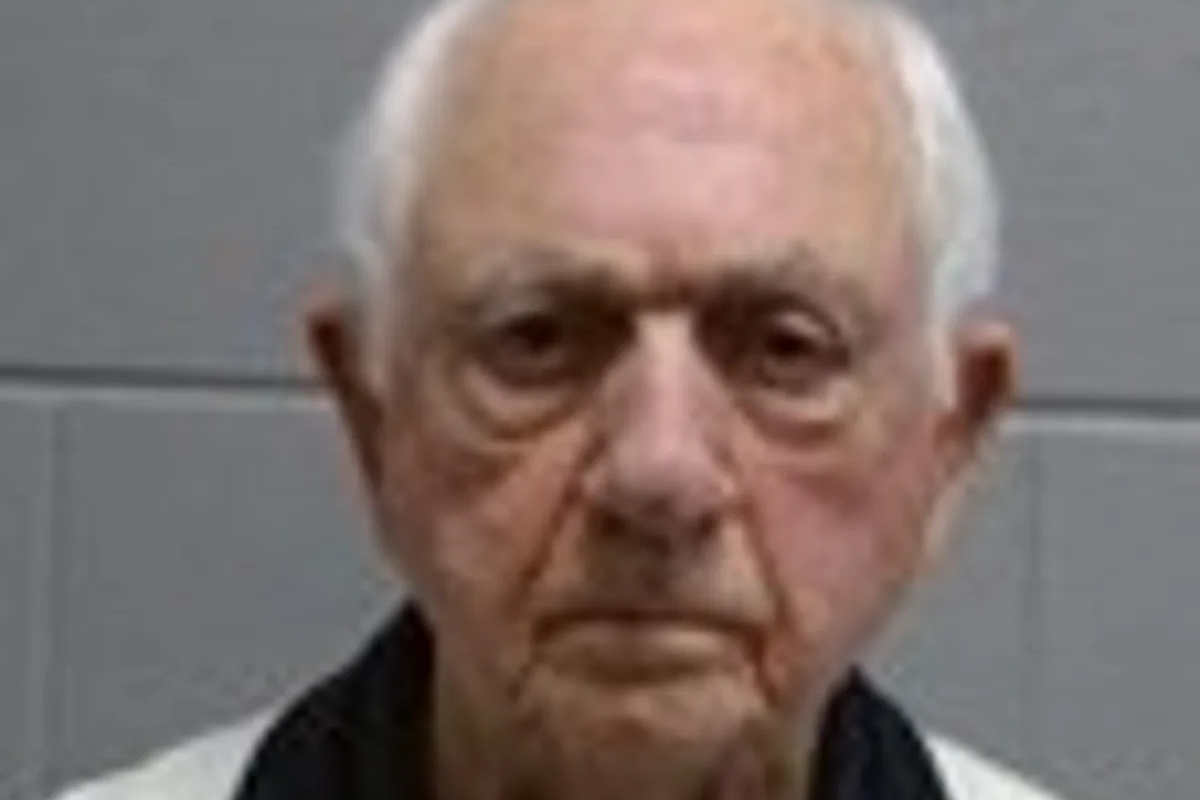 Close-up of an elderly man with short white hair and a neutral, serious expression against a gray wall backdrop.