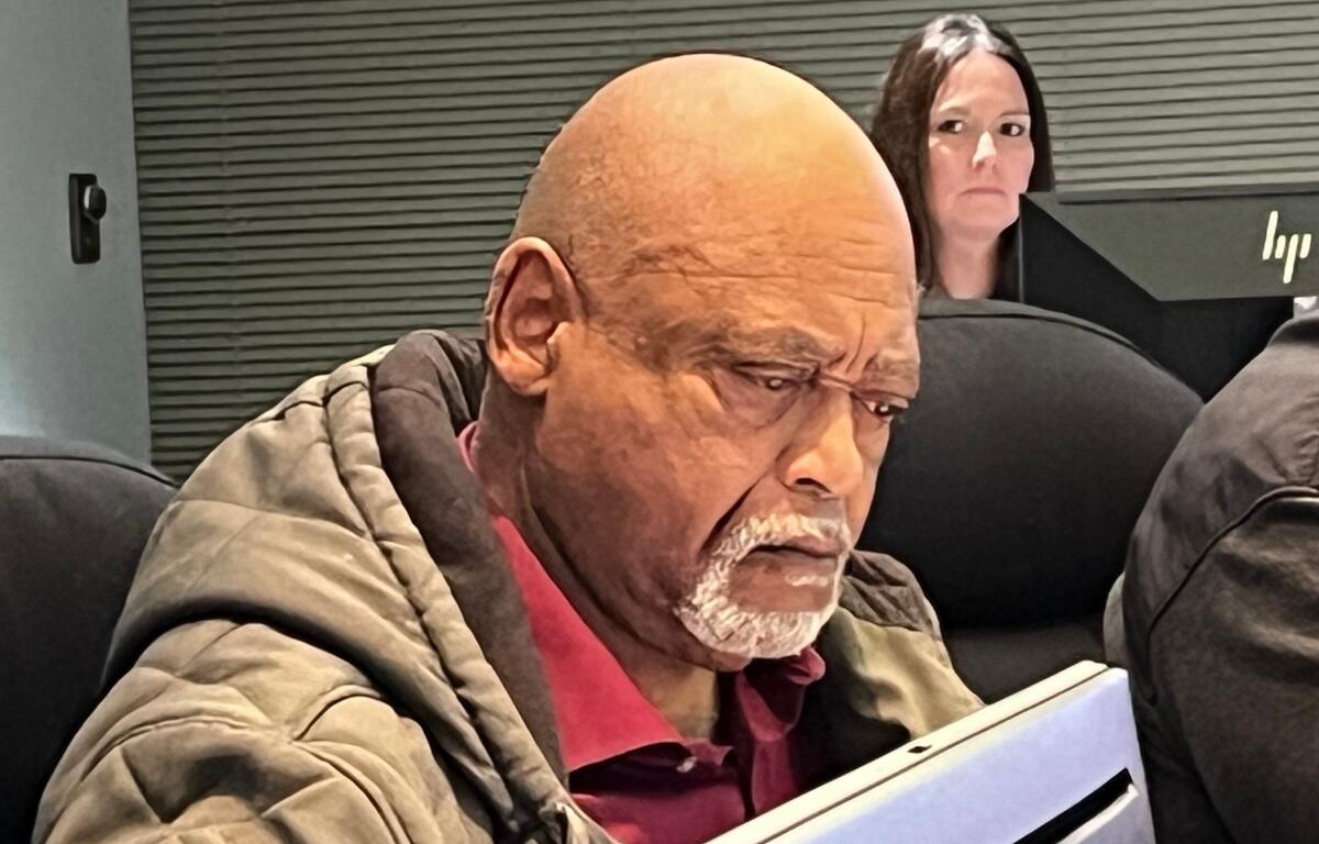 Man with a bald head and gray beard reading a tablet in an office setting, with a woman and an HP monitor in the background.