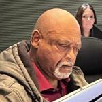 Man with a bald head and gray beard reading a tablet in an office setting, with a woman and an HP monitor in the background.