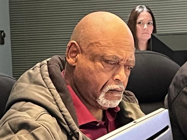 Man with a bald head and gray beard reading a tablet in an office setting, with a woman and an HP monitor in the background.
