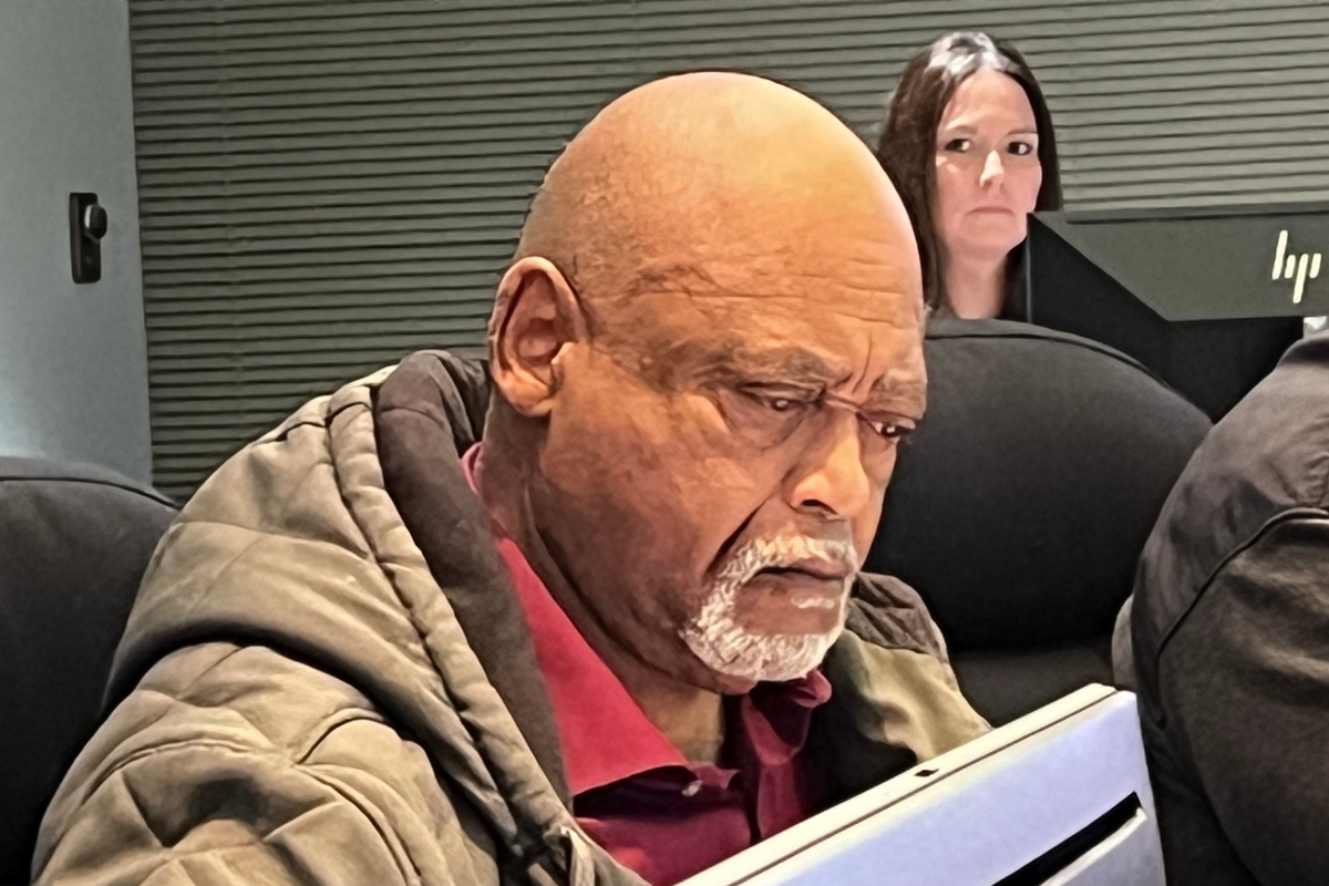 Man with a bald head and gray beard reading a tablet in an office setting, with a woman and an HP monitor in the background.