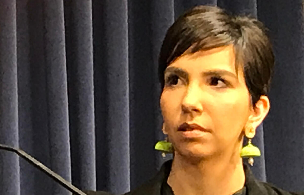 Woman with short dark hair and lime-green earrings speaks on a stage in front of dark curtains, looking to the side.