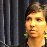 Woman with short dark hair and lime-green earrings speaks on a stage in front of dark curtains, looking to the side.