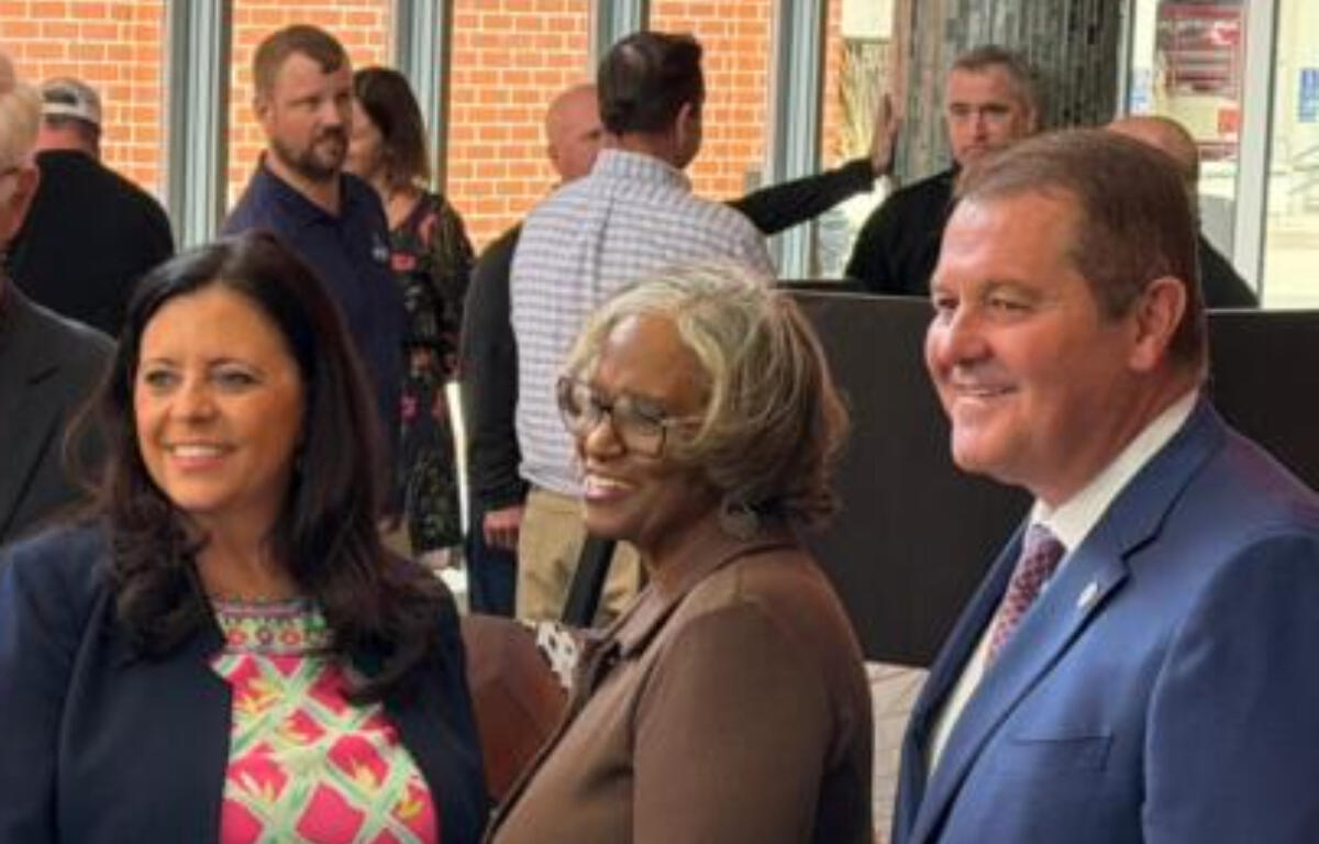 Three adults posing and smiling at a social event, with two women in front and a man in a blue suit on the right, inside a lobby area with other guests in the background.