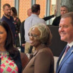 Three adults posing and smiling at a social event, with two women in front and a man in a blue suit on the right, inside a lobby area with other guests in the background.