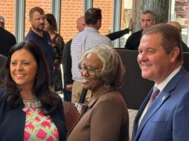 Three adults posing and smiling at a social event, with two women in front and a man in a blue suit on the right, inside a lobby area with other guests in the background.