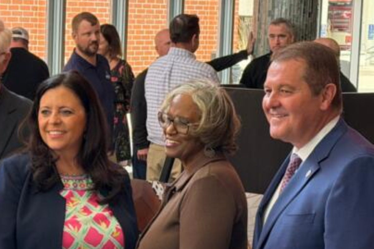 Three adults posing and smiling at a social event, with two women in front and a man in a blue suit on the right, inside a lobby area with other guests in the background.