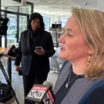 Side-profile of a blonde woman in a dark blue top speaking to reporters, microphones labeled Capitol City Now in front of her.