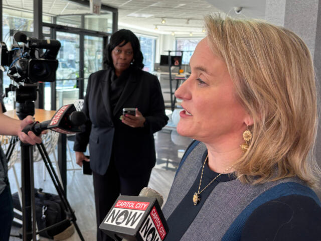 Side-profile of a blonde woman in a dark blue top speaking to reporters, microphones labeled Capitol City Now in front of her.