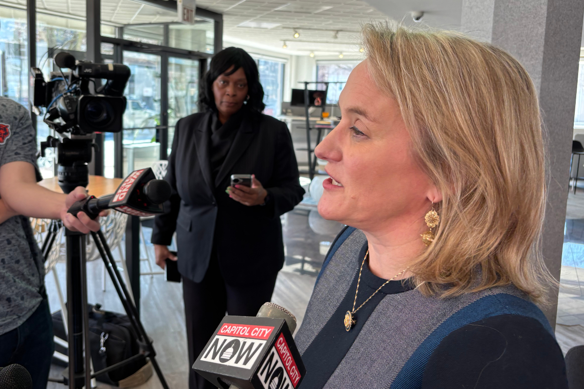 Side-profile of a blonde woman in a dark blue top speaking to reporters, microphones labeled Capitol City Now in front of her.