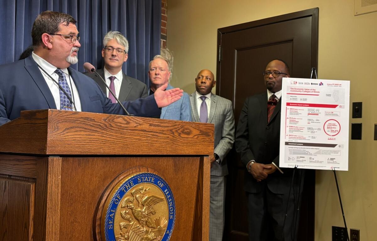 Speaker in a dark suit talks at a wooden podium with the state seal; four men in suits stand behind him, with a large informational poster on a stand nearby.