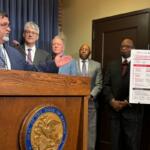Speaker in a dark suit talks at a wooden podium with the state seal; four men in suits stand behind him, with a large informational poster on a stand nearby.
