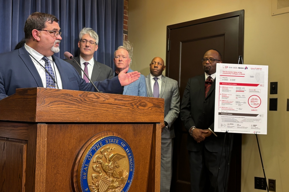 Speaker in a dark suit talks at a wooden podium with the state seal; four men in suits stand behind him, with a large informational poster on a stand nearby.