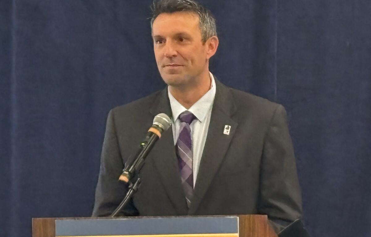 Man in a dark suit and purple striped tie speaks at a lectern with a microphone.