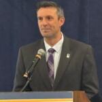 Man in a dark suit and purple striped tie speaks at a lectern with a microphone.