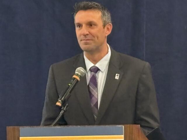 Man in a dark suit and purple striped tie speaks at a lectern with a microphone.