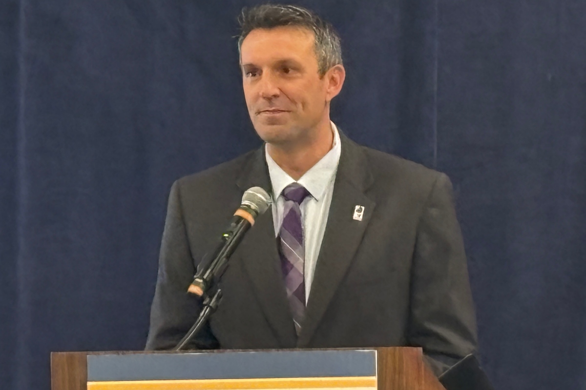 Man in a dark suit and purple striped tie speaks at a lectern with a microphone.