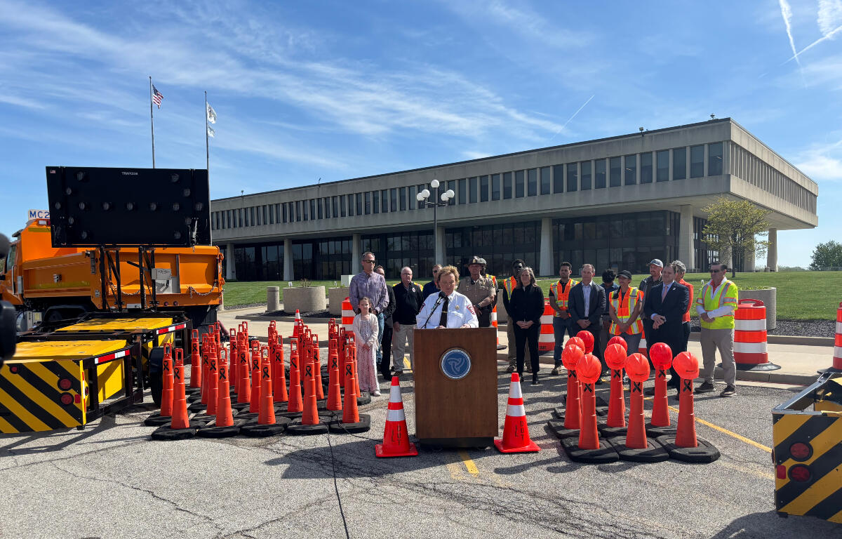 Officials hold an outdoor press conference behind a podium surrounded by orange traffic cones and a large screen on a truck nearby. Nearby, people in safety vests and formal attire stand in front of a modern office building.