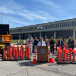 Officials hold an outdoor press conference behind a podium surrounded by orange traffic cones and a large screen on a truck nearby. Nearby, people in safety vests and formal attire stand in front of a modern office building.