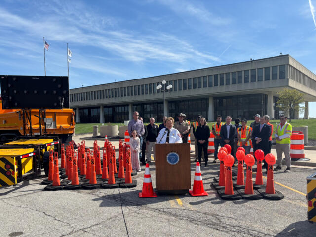 Officials hold an outdoor press conference behind a podium surrounded by orange traffic cones and a large screen on a truck nearby. Nearby, people in safety vests and formal attire stand in front of a modern office building.