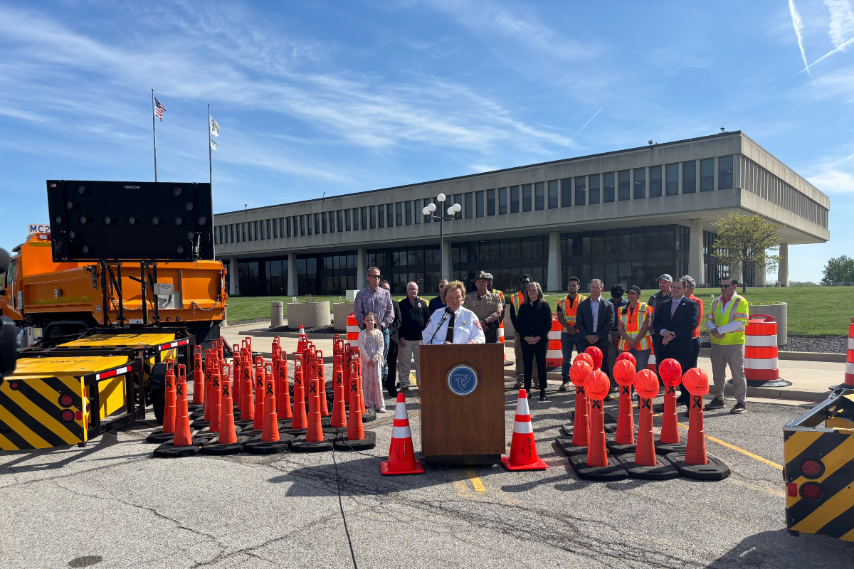 Officials hold an outdoor press conference behind a podium surrounded by orange traffic cones and a large screen on a truck nearby. Nearby, people in safety vests and formal attire stand in front of a modern office building.
