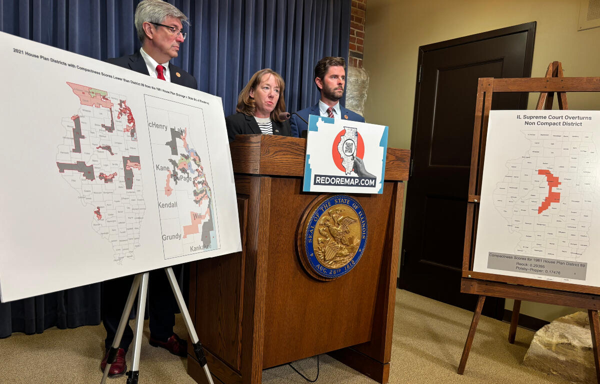 Three people stand behind a wooden podium with a REDOREMAP.COM sign, flanked by large district maps on easels.