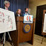 Three people stand behind a wooden podium with a REDOREMAP.COM sign, flanked by large district maps on easels.