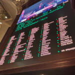 Inside an ornate hall, a large electronic scoreboard lists names in columns with green and red markers, with a TV feed above.