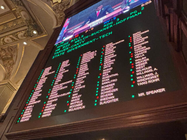 Inside an ornate hall, a large electronic scoreboard lists names in columns with green and red markers, with a TV feed above.