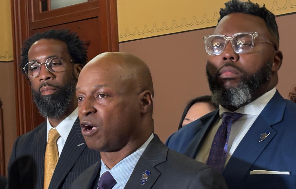 Three men in formal suits at a indoors formal event, one speaking while others stand nearby, serious expressions behind a wooden door and decorative wall.