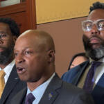 Three men in formal suits at a indoors formal event, one speaking while others stand nearby, serious expressions behind a wooden door and decorative wall.