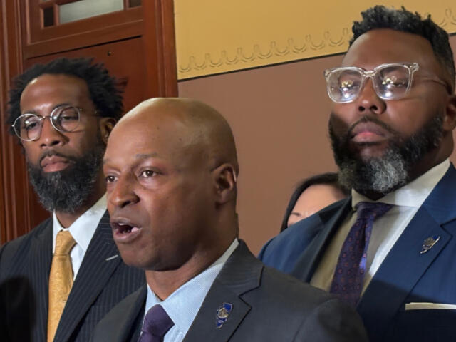 Three men in formal suits at a indoors formal event, one speaking while others stand nearby, serious expressions behind a wooden door and decorative wall.
