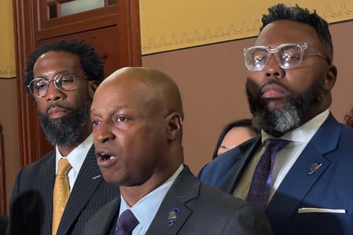 Three men in formal suits at a indoors formal event, one speaking while others stand nearby, serious expressions behind a wooden door and decorative wall.