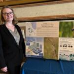 Woman in a black blazer stands beside a multi-panel poster board about marsh bird habitats.