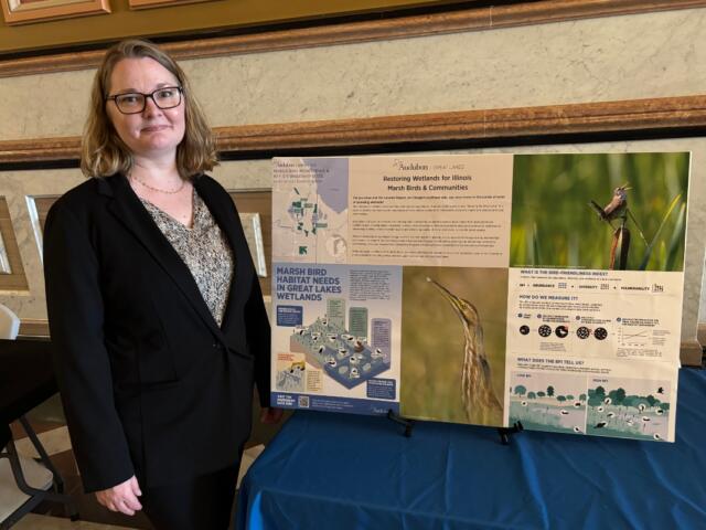 Woman in a black blazer stands beside a multi-panel poster board about marsh bird habitats.