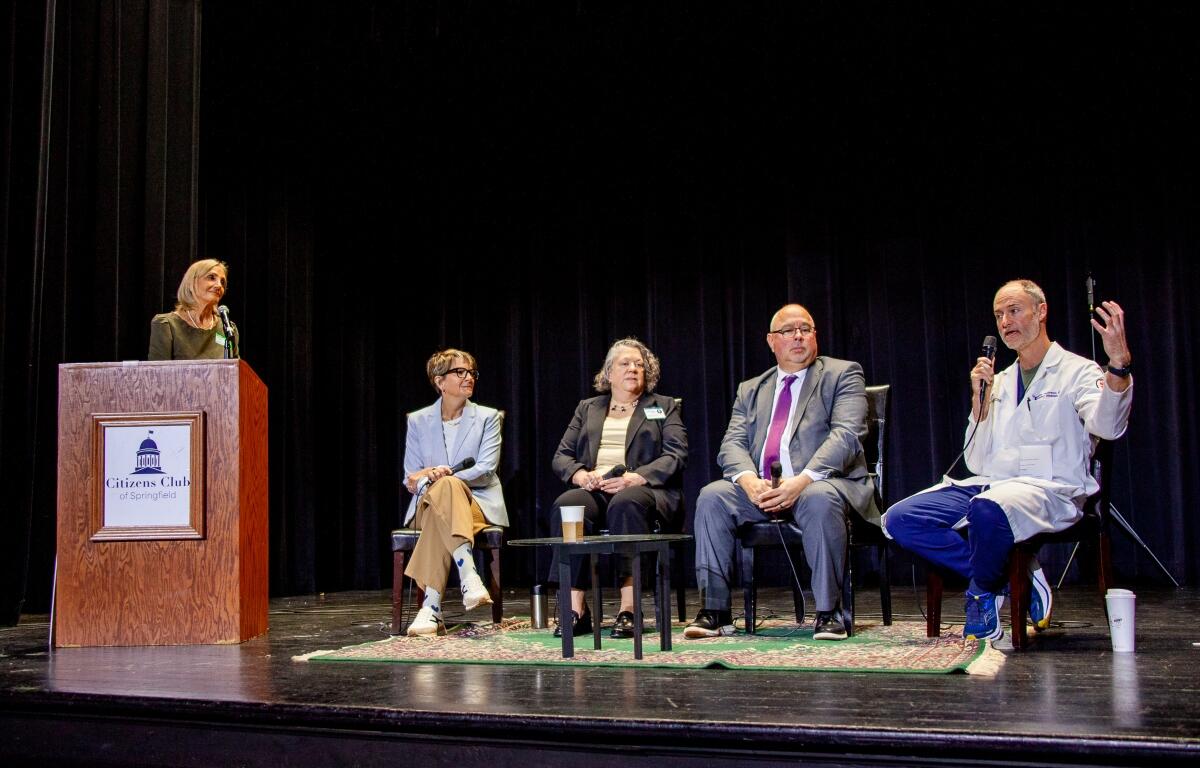 Woman at a podium speaking into a microphone on stage; panelists seated behind her with coffee cup on table and microphones present.