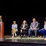 Woman at a podium speaking into a microphone on stage; panelists seated behind her with coffee cup on table and microphones present.
