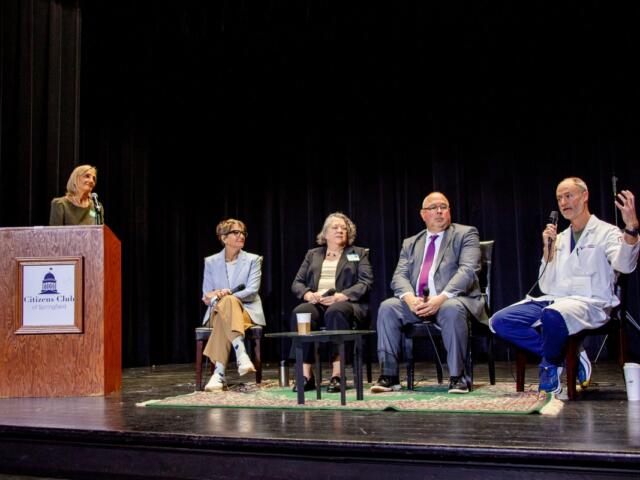 Woman at a podium speaking into a microphone on stage; panelists seated behind her with coffee cup on table and microphones present.