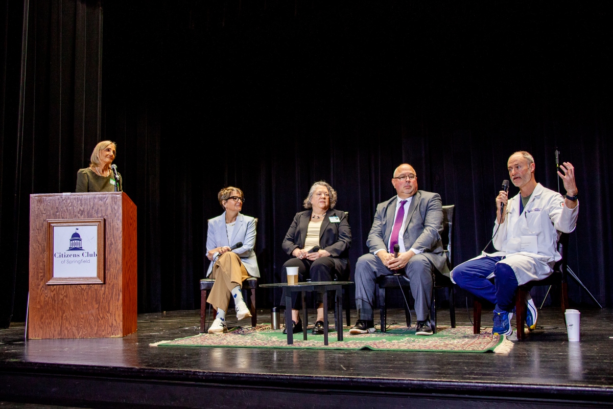 Woman at a podium speaking into a microphone on stage; panelists seated behind her with coffee cup on table and microphones present.