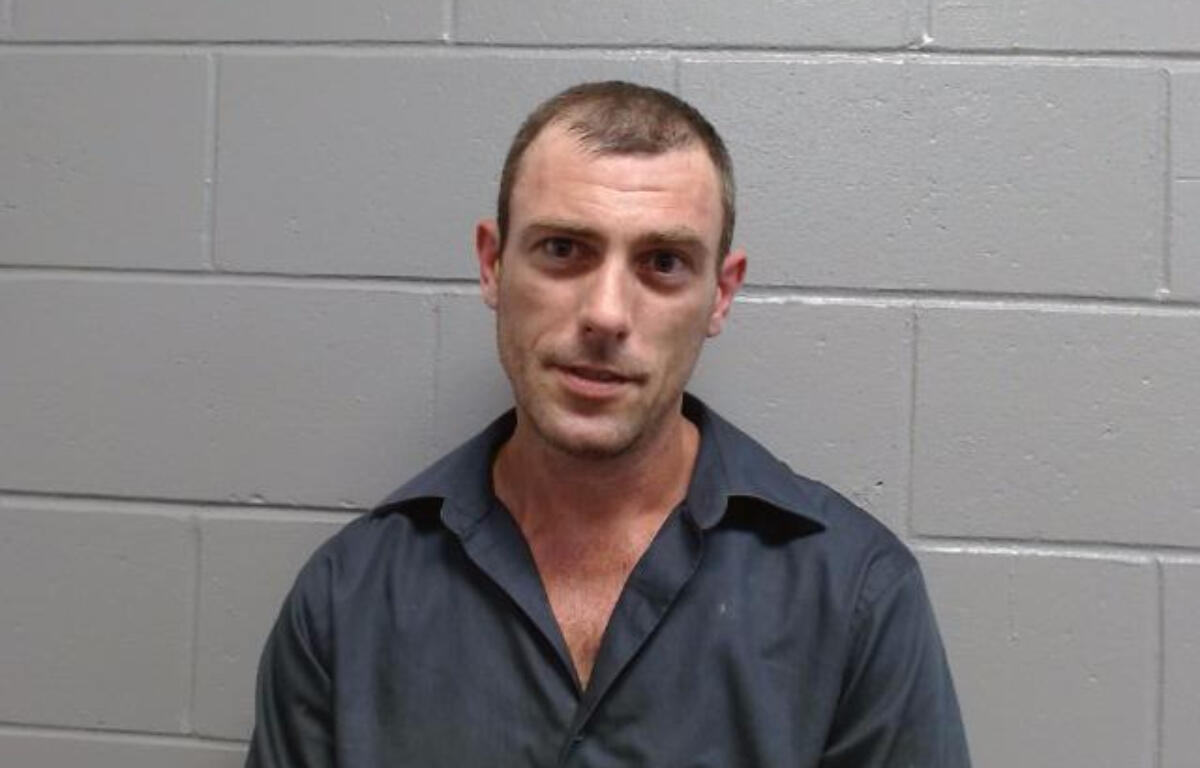 Mugshot-style photo of a man in a dark shirt standing against a gray cinder block wall.